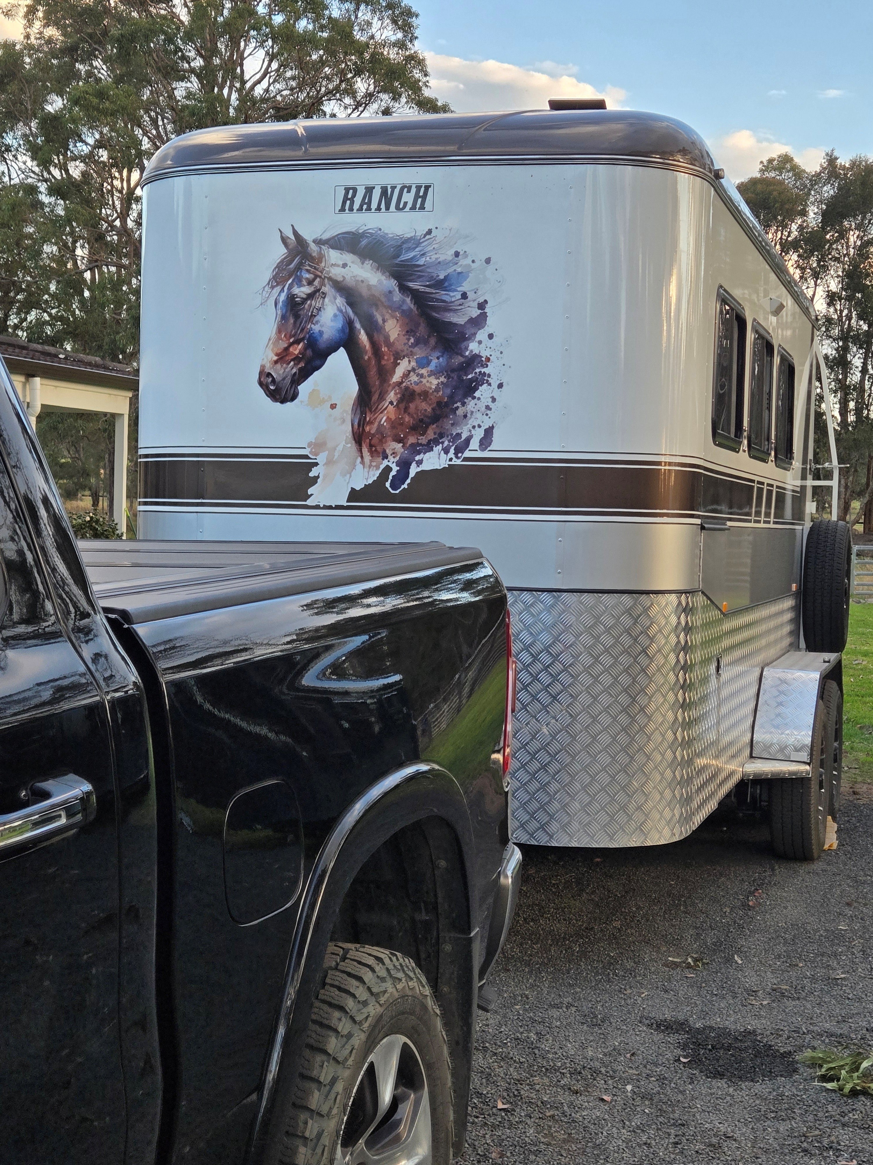 A silver horse trailer with a large horse graphic and the word 'RANCH' on the side, parked outdoors next to a black pickup truck on a gravel surface.