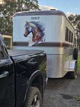 A silver horse trailer with a large horse graphic and the word 'RANCH' on the side, parked outdoors next to a black pickup truck on a gravel surface.