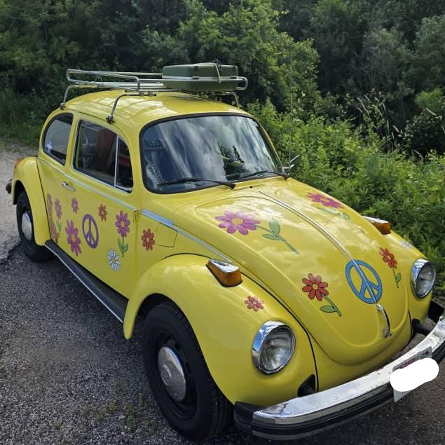 Yellow vintage car with floral and peace symbol design parked on a road surrounded by trees.