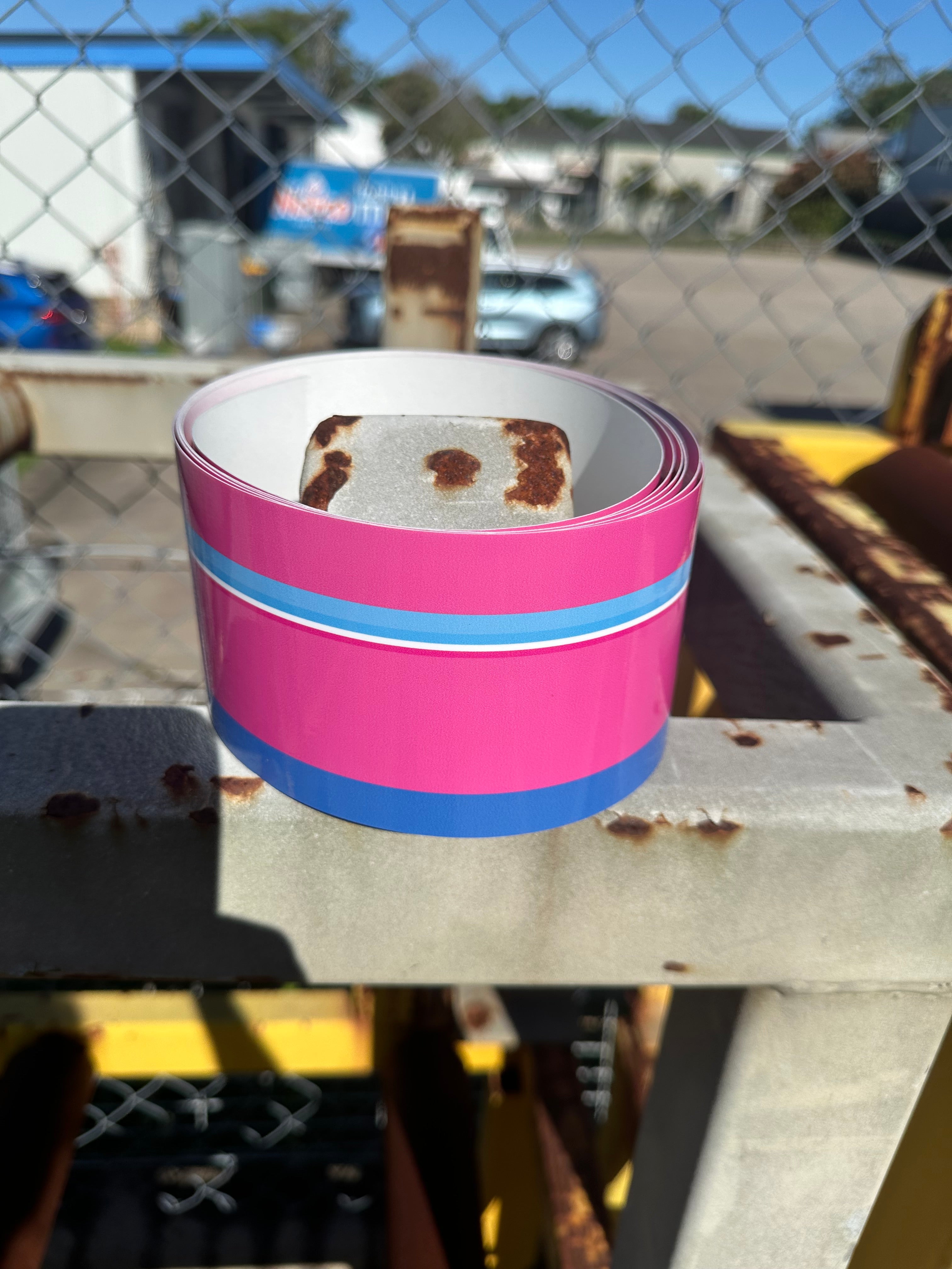 Pink and blue tape roll attached to a metal surface with a chain-link fence and building in the background.